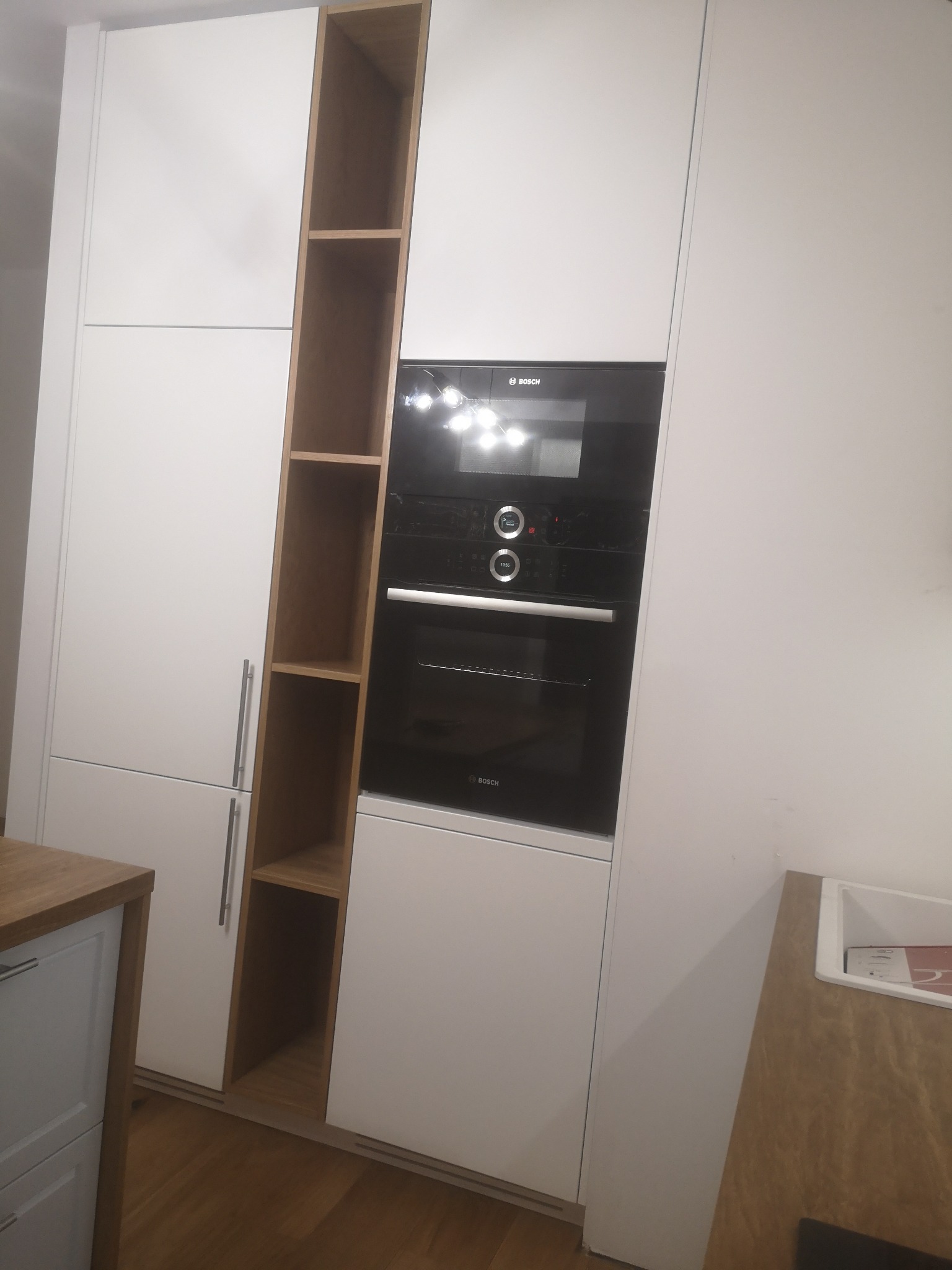 Biała zabudowa kuchenna z piekarnikiem i otwartym regałem z jasnego drewna, widoczne metalowe uchwyty i fragment blatu.