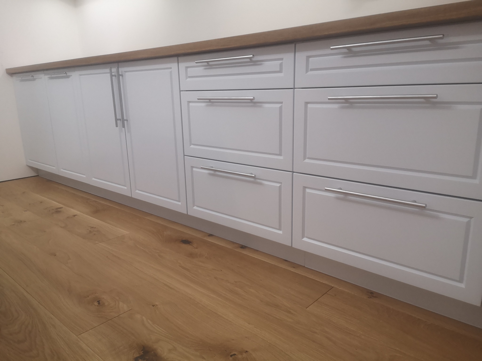 Szare szafki kuchenne z drewnianym blatem i podłogą z paneli w ciepłym odcieniu, widok z niskiej perspektywy.