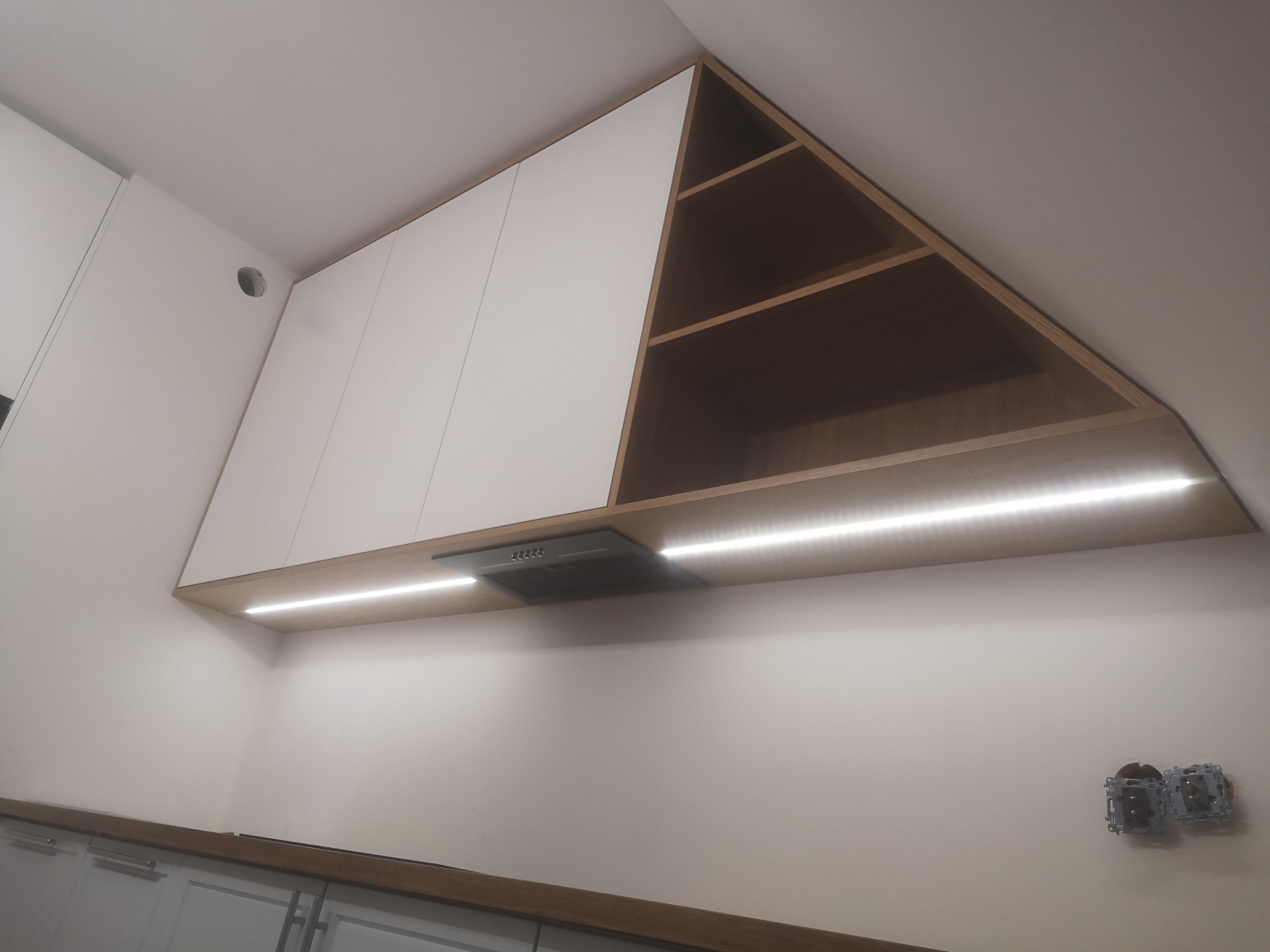 Białe szafki kuchenne pod skosem sufitu z oświetleniem LED, szary okap i widoczne gniazdka elektryczne na ścianie.