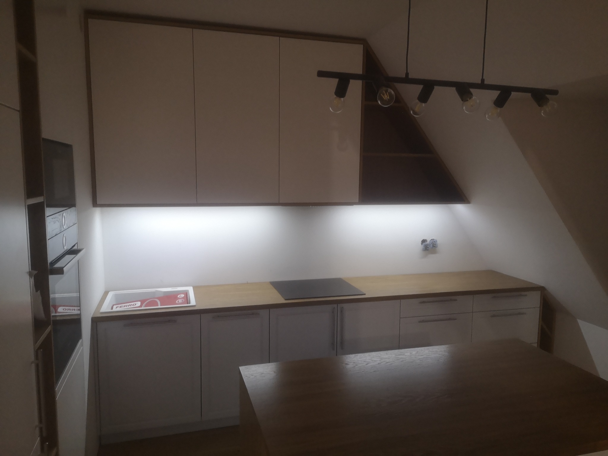 Kuchnia w skosie z białymi szafkami, drewnianym blatem i czarną lampą sufitową z żarówkami edisona, widoczny zlewozmywak z opakowaniem Ferro.