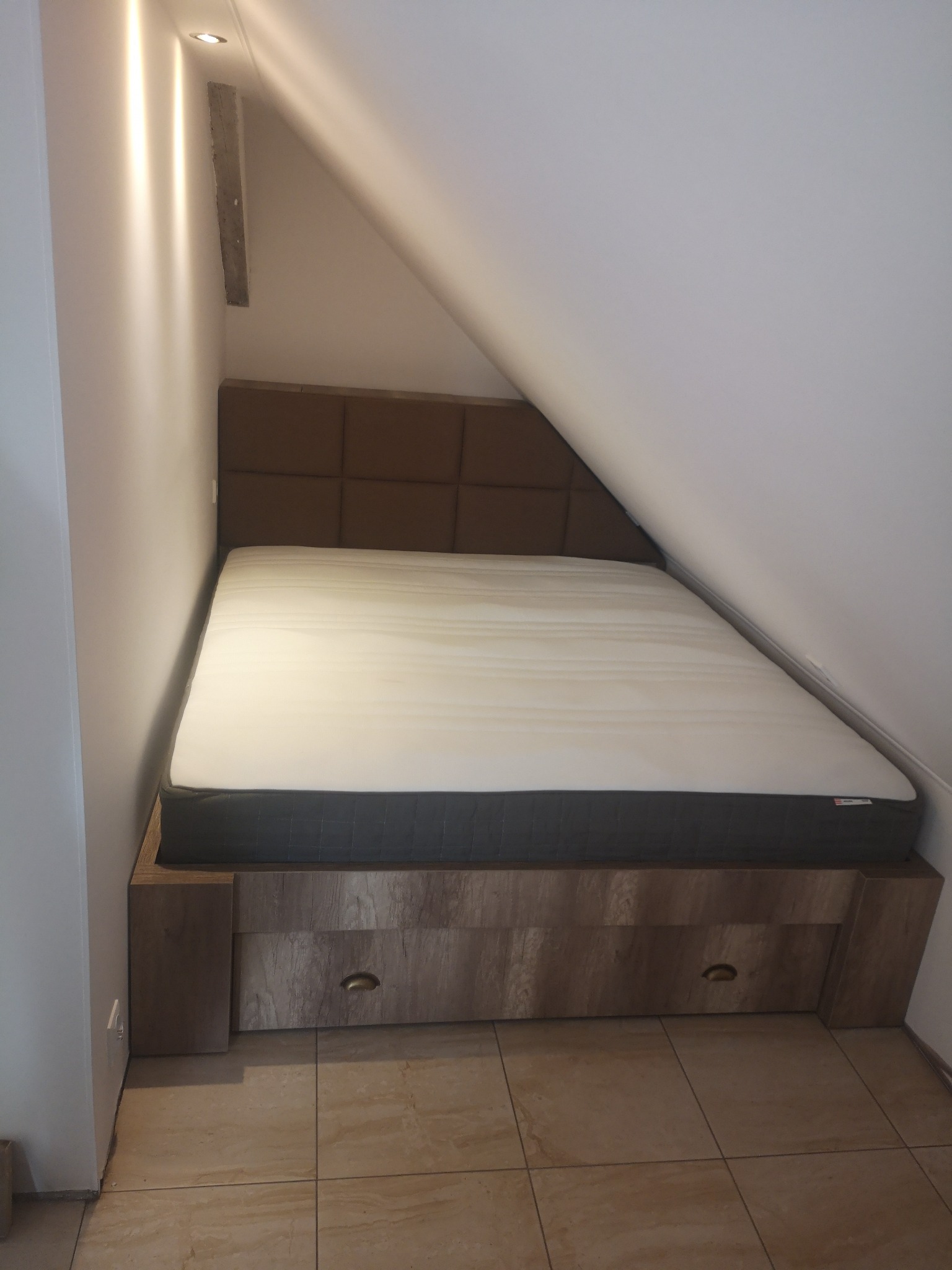 Łóżko z tapicerowanym zagłówkiem i szufladami wbudowane w skos poddasza, wykończone drewnopodobną okleiną, na podłodze ceramiczne płytki imitujące kamień.