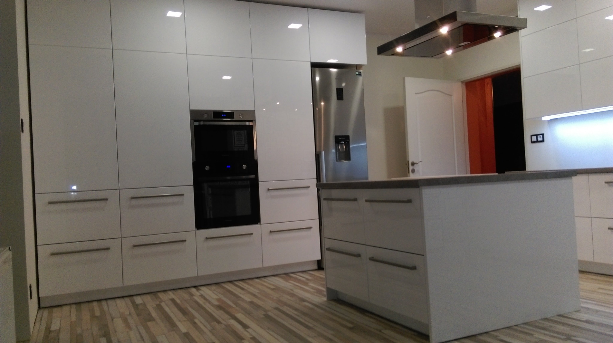 Nowoczesna, biała kuchnia z połyskiem, wyposażona w zabudowany piekarnik, lodówkę side-by-side i wyspę kuchenną z szarym blatem, na podłodze panele imitujące drewno.