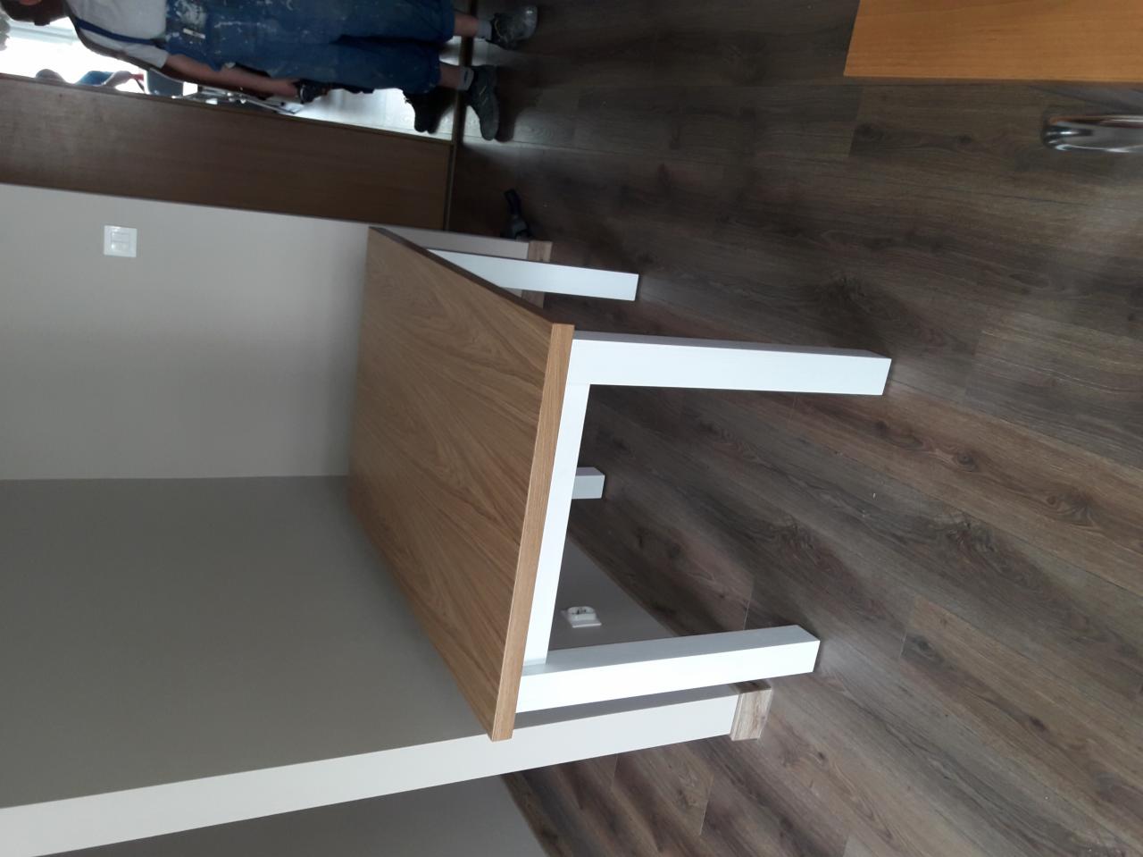 Montaż blatu z drewna i białych nóg w szafie wnękowej, widoczny fragment sylwetki montera w ubraniu roboczym.