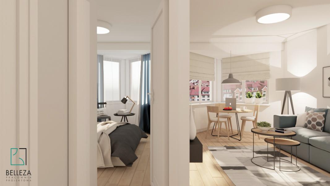 Wnętrze mieszkania z widokiem na sypialnię i salon z jadalnią, jasne ściany, drewniana podłoga, okna z roletami rzymskimi i zasłonami, nowoczesne meble i oświetlenie, logo firmy w lewym dolnym rogu.