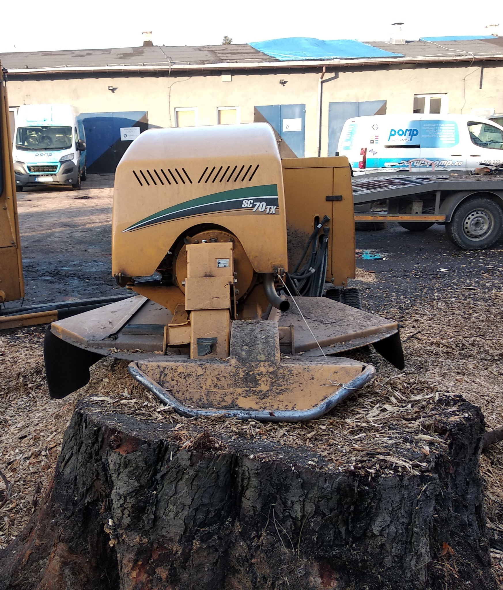 Frezarka do pni SC70TX na pniu drzewa, otoczona wiórami, na tle budynków gospodarczych i samochodów dostawczych.