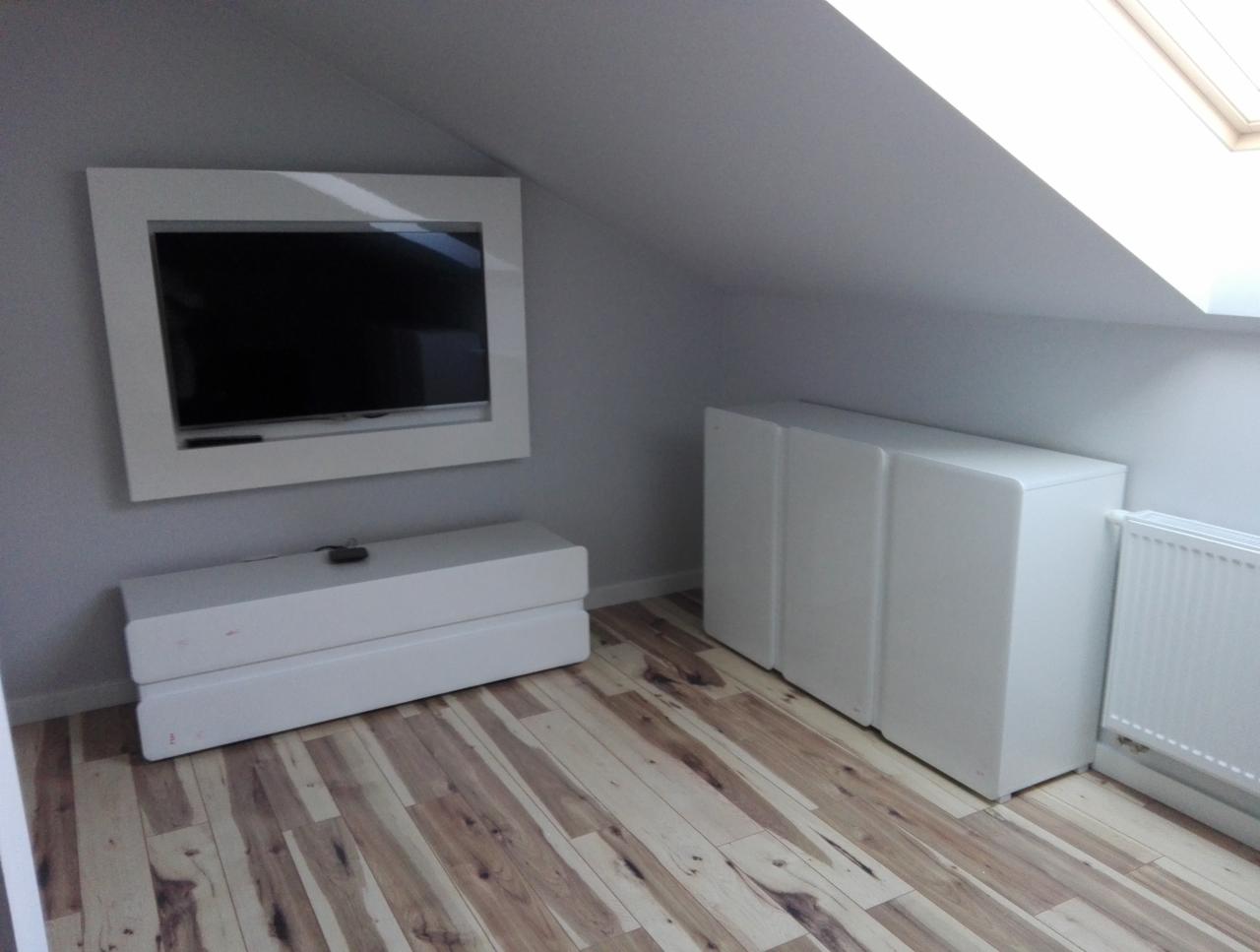 Białe szafki i szafka RTV pod telewizorem w ramie, na poddaszu ze skośnym sufitem i oknem dachowym.