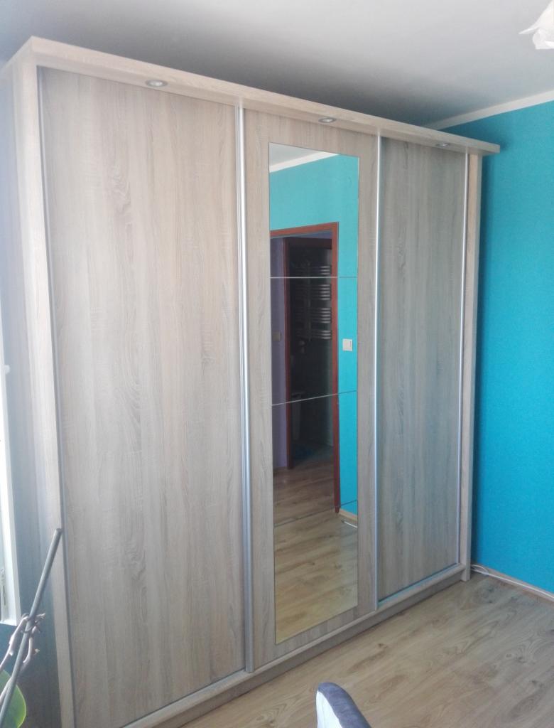 Szafa przesuwna z lustrem na środkowych drzwiach, wykończona okleiną w kolorze jasnego drewna, widoczna na tle turkusowej ściany i drewnianej podłogi.