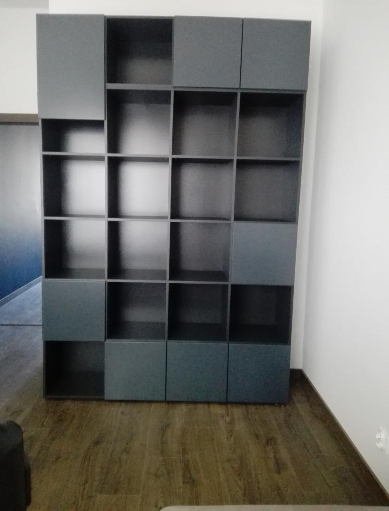 Szara, modułowa biblioteczka z kwadratowymi półkami, wykonana na wymiar, z widocznym fragmentem drewnianej podłogi i białej ściany.