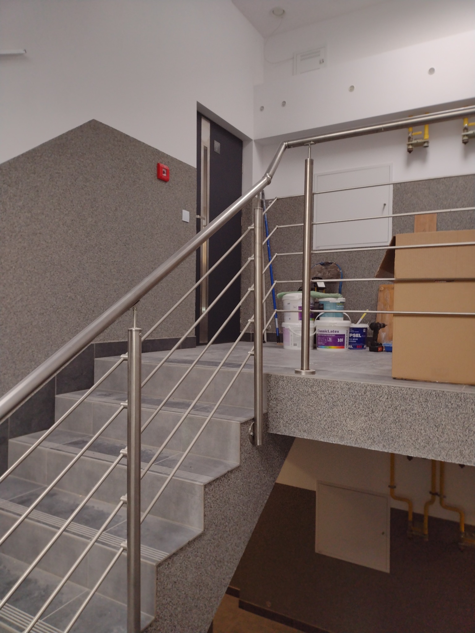 Klatka schodowa w trakcie remontu z metalową balustradą, widoczne wiadra z farbą, kartony i narzędzia, ściany częściowo wykończone.