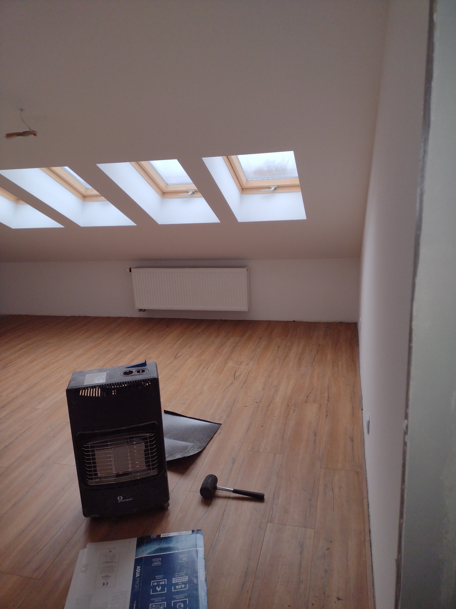 Pokój na poddaszu z trzema oknami dachowymi, panelami podłogowymi, grzejnikiem, przenośnym piecykiem grzewczym, gumowym młotkiem i opakowaniem po panelach.