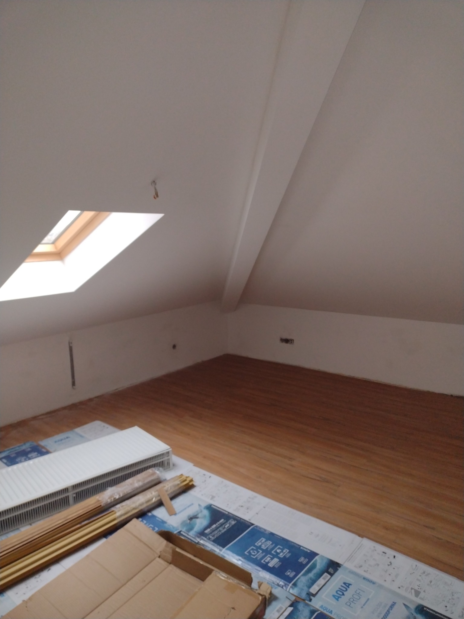 Poddasze po remoncie z drewnianą podłogą, okno dachowe i grzejnik na podłodze na opakowaniach paneli.
