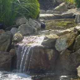Oczko wodne z kaskadą wodną, otoczone kamieniami i roślinnością ozdobną, z widocznym elementem systemu filtracji w tle.