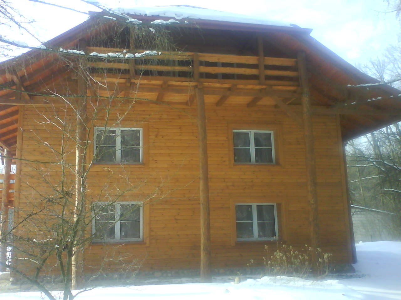 Dwupiętrowy dom z drewna, z balkonem na drugim piętrze, widoczny w zimowej scenerii z drzewami w tle.