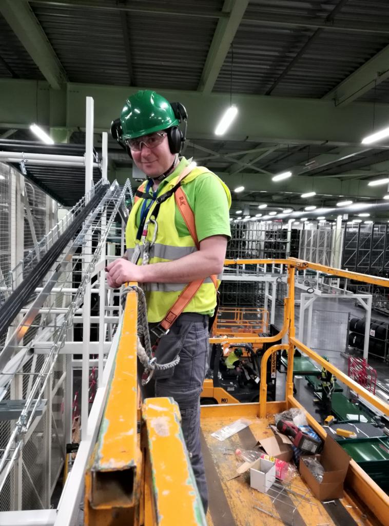 Pracownik w zielonym kasku i okularach ochronnych, stojący na podnośniku koszowym, w tle instalacja przewodów w hali przemysłowej.