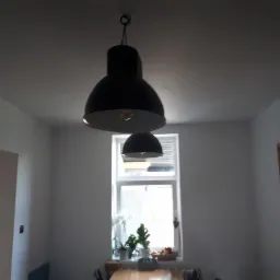 Wnętrze pokoju z dwoma czarnymi lampami wiszącymi nad stołem, okno z białymi roletami i roślinami na parapecie, białe ściany i sufit.