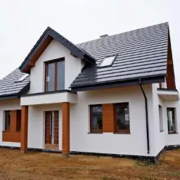 Nowoczesny, dwukondygnacyjny dom jednorodzinny z ciemną dachówką, białymi ścianami i drewnianymi akcentami, w trakcie wykończenia terenu wokół budynku.