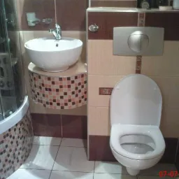 Wykończona łazienka z kabiną prysznicową, umywalką na szafce z mozaiką oraz toaletą wiszącą z przyciskiem spłukującym na ścianie wyłożonej płytkami w odcieniach beżu i brązu.