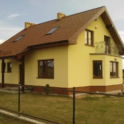 Żółty dom murowany z brązowym dachem i oknami, widok z zewnątrz na elewację z balkonem z ozdobną balustradą.