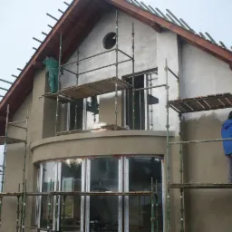 Dwóch pracowników na rusztowaniu podczas prac elewacyjnych na dwupiętrowym domu z okrągłym oknem na poddaszu, widoczne świeżo nałożone tynki i przygotowane drewniane elementy dachu.