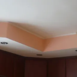 Wykończony sufit z oświetleniem punktowym i fragmentem lampy, nad brązowymi szafkami kuchennymi, z pomalowaną na brzoskwiniowo wnęką.