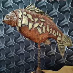 Arkadi - Ręcznie wykonana metalowa rzeźba karpia w kolorach miedzi i złota, zamontowana na postumencie z przekładni, na tle wzorzystej tapety.