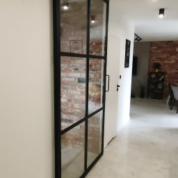 Arkadi - Szklane drzwi przesuwne z czarną metalową ramą w stylu industrialnym, dzielone na prostokątne segmenty, zamontowane w jasnym korytarzu z widoczną ceglaną ścianą w tle.
