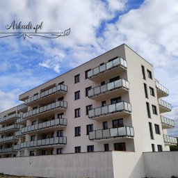 Arkadi - Nowoczesny budynek mieszkalny z balkonami zabezpieczonymi szklanymi balustradami, widok z dołu na tle błękitnego nieba z chmurami, w lewym górnym rogu logo firmy Arkadi.pl.