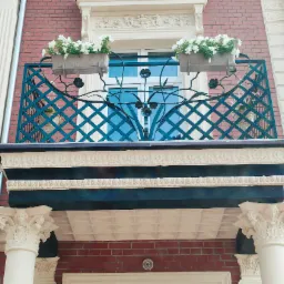 Elegancki balkon z ozdobną, ciemnozieloną balustradą z motywem kwiatowym, ozdobiony skrzynkami z białymi kwiatami, wsparty na kolumnach z dekoracyjnymi kapitelami, na tle ceglanej ściany.