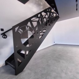 Arkadi - Nowoczesne schody metalowe z ażurową balustradą, na której wycięto logo BMW i napis MPOWER. Całość w czarnym kolorze, na tle białych ścian i szarej podłogi.