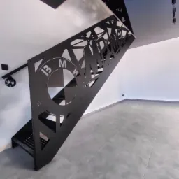 Nowoczesne schody metalowe z ażurową balustradą, na której wycięto logo BMW i napis MPOWER. Całość w czarnym kolorze, na tle białych ścian i szarej podłogi.