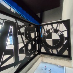Arkadi - Czarna, metalowa balustrada schodowa z wyciętymi laserowo wzorami geometrycznymi i logo BMW, widok z góry na schody w trakcie wykończenia.