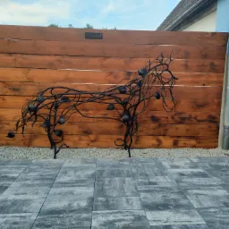 Kuta, metalowa rzeźba konia z liśćmi i owocami na tle drewnianego ogrodzenia z tabliczką 'Arkadi.pl', ułożona na szarej kostce brukowej.