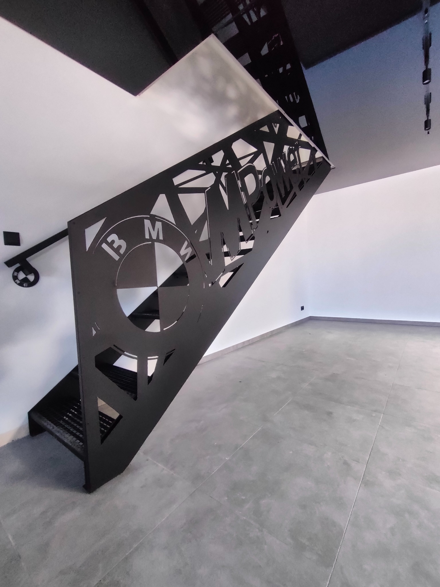 Nowoczesne schody metalowe z ażurową balustradą, na której wycięto logo BMW i napis MPOWER. Całość w czarnym kolorze, na tle białych ścian i szarej podłogi.