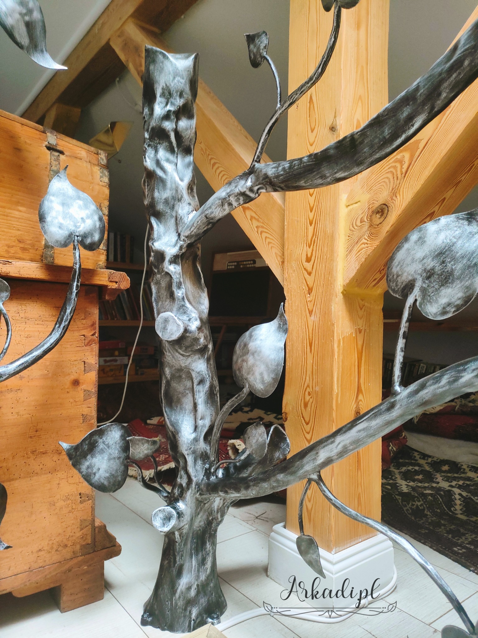 Metalowa balustrada wewnętrzna w kształcie drzewa z liśćmi, wsparta na drewnianej kolumnie w rustykalnym wnętrzu.