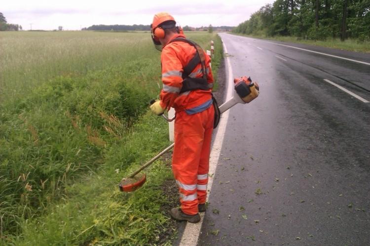Pracownik w pomarańczowym kombinezonie i ochronnikach słuchu, przy użyciu kosy spalinowej, przycina trawę wzdłuż mokrej asfaltowej drogi, z polem w tle.