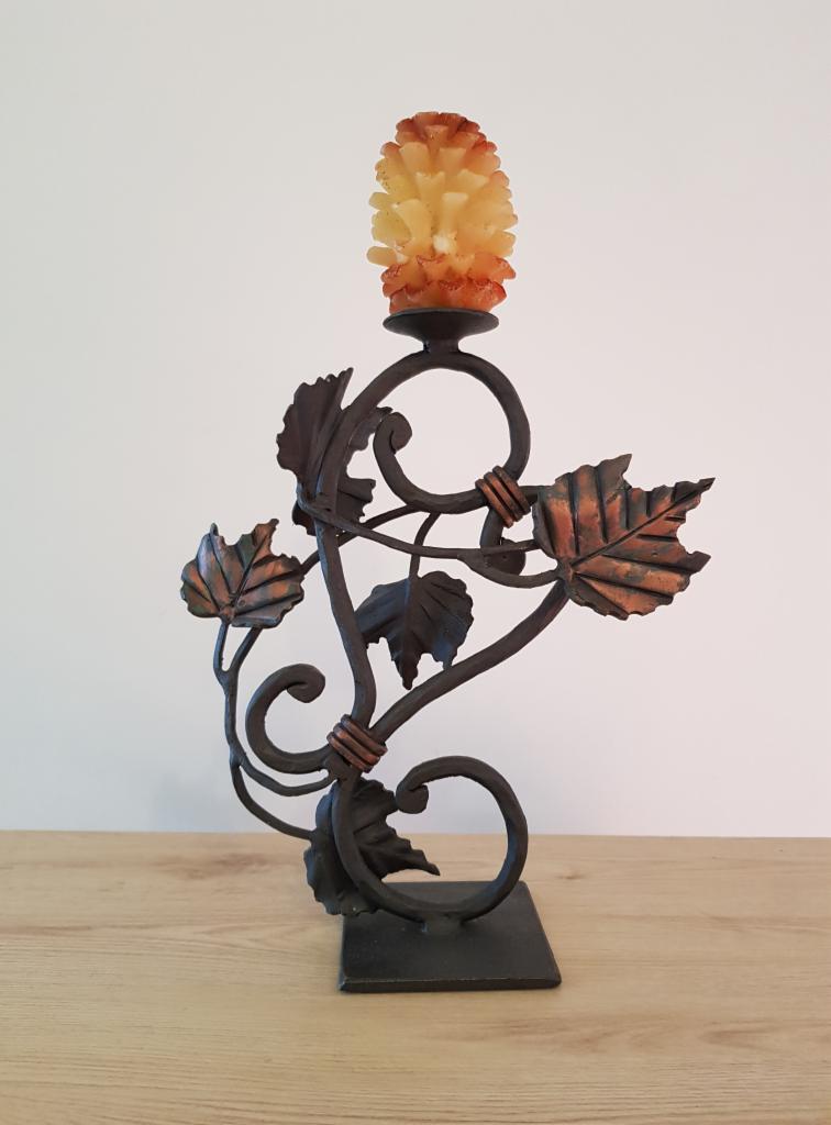 Kuty świecznik z metalu, zdobiony motywem liści winogron i świecą w kształcie szyszki, stojący na drewnianym blacie.