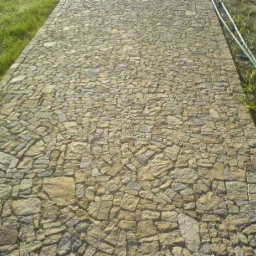 Ułożona ścieżka z nieregularnych, jasnych kostek granitowych, widok z góry, obok trawa i zielony wąż ogrodowy.