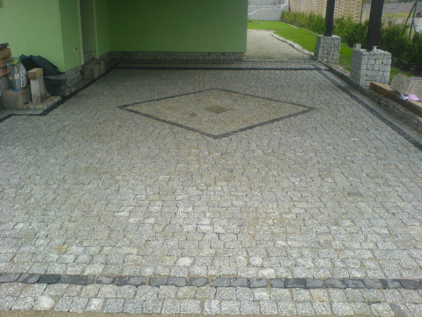 Ułożona nawierzchnia z kostki granitowej w odcieniach szarości, z dekoracyjnym wzorem rombu w centralnej części, obrzeża wykończone ciemniejszą kostką, widoczne elementy budynku w tle.
