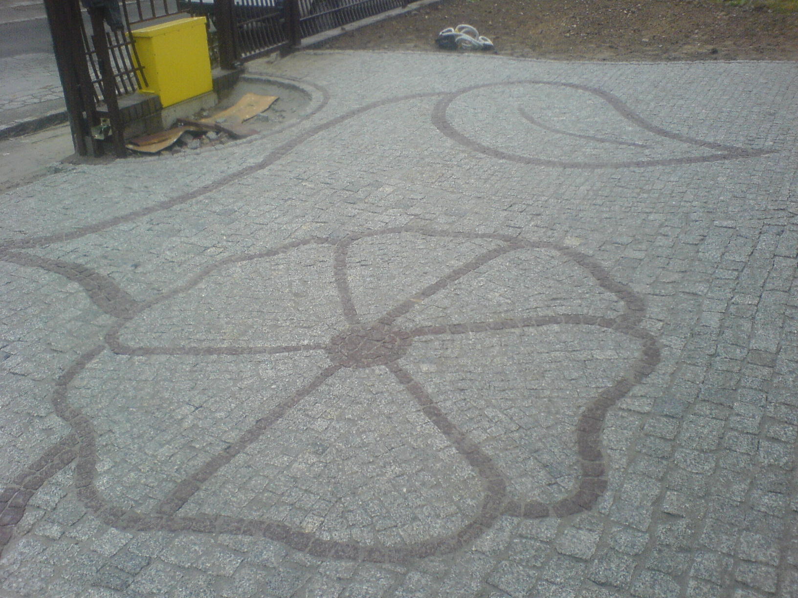 Ułożona kostka brukowa w szarym odcieniu, z wzorem kwiatu i liścia wykonanym z kostki w ciemniejszym odcieniu, widoczne elementy infrastruktury miejskiej w tle.