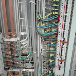 Wnętrze szafy elektrycznej z uporządkowanymi kablami w różnych kolorach, widoczne metalowe szyny i listwy zaciskowe.