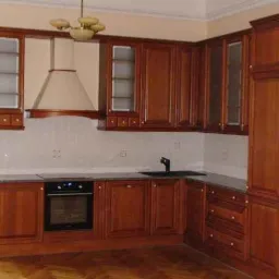 Klasyczna kuchnia z drewnianymi szafkami w odcieniach brązu, szarym blatem i białymi płytkami, oświetlona lampą w stylu retro.