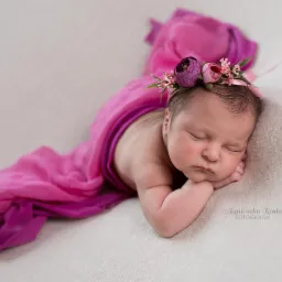Śpiąca noworodka owinięta w różowy materiał, z wiankiem z kwiatów na głowie, delikatne oświetlenie.