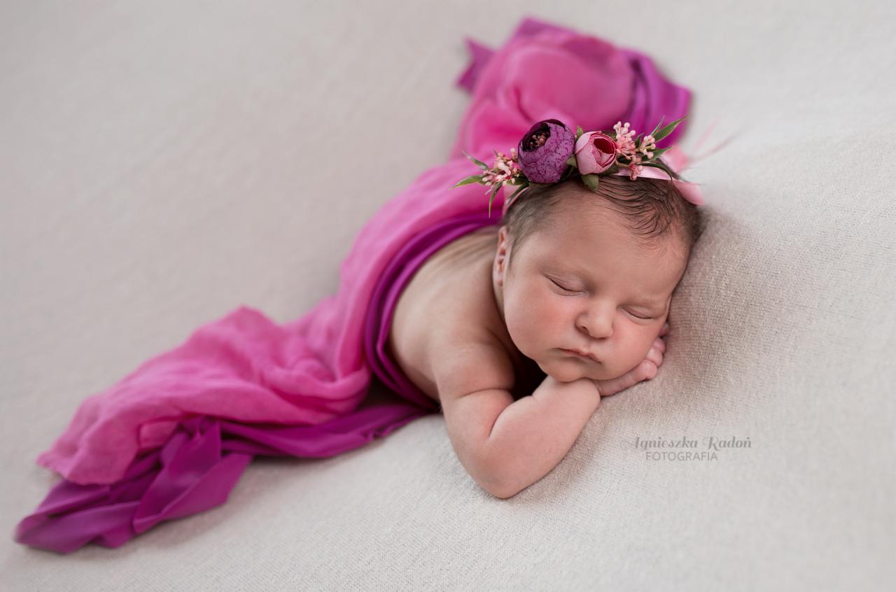 Śpiąca noworodka owinięta w różowy materiał, z wiankiem z kwiatów na głowie, delikatne oświetlenie.