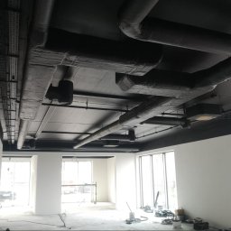 Maestro usługi remontowo- wykończeniowe Tomasz Serocki - Pomieszczenie biurowe po malowaniu sufitu na czarno, widoczne rury wentylacyjne i instalacje. Ściany pomalowane na biało, duże okna w tle.