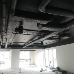 Pomieszczenie biurowe po malowaniu sufitu na czarno, widoczne rury wentylacyjne i instalacje. Ściany pomalowane na biało, duże okna w tle.