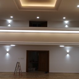 Maestro usługi remontowo- wykończeniowe Tomasz Serocki - Jasne, przestronne wnętrze po remoncie z oświetleniem punktowym, drewnianą podłogą i schodami. Widoczna drabina sugeruje prace wykończeniowe.