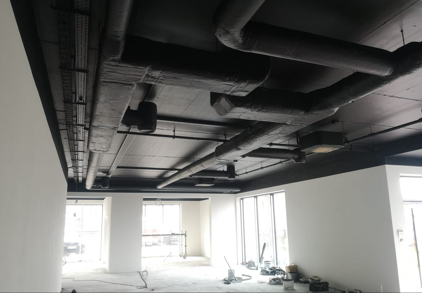 Pomieszczenie biurowe po malowaniu sufitu na czarno, widoczne rury wentylacyjne i instalacje. Ściany pomalowane na biało, duże okna w tle.