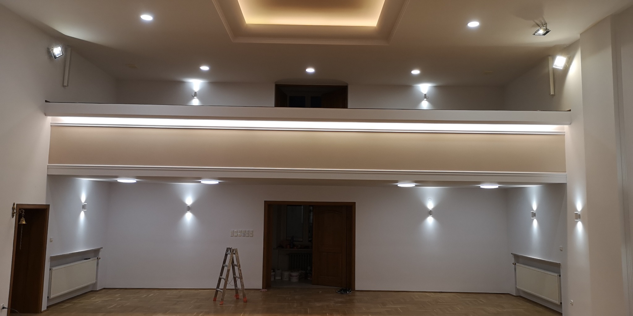 Jasne, przestronne wnętrze po remoncie z oświetleniem punktowym, drewnianą podłogą i schodami. Widoczna drabina sugeruje prace wykończeniowe.