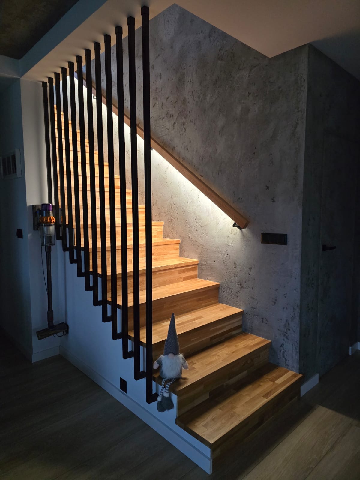 Nowoczesne schody drewniane z czarną balustradą i oświetleniem LED, ściana z efektem betonu. Na schodach siedzi dekoracyjny skrzat.