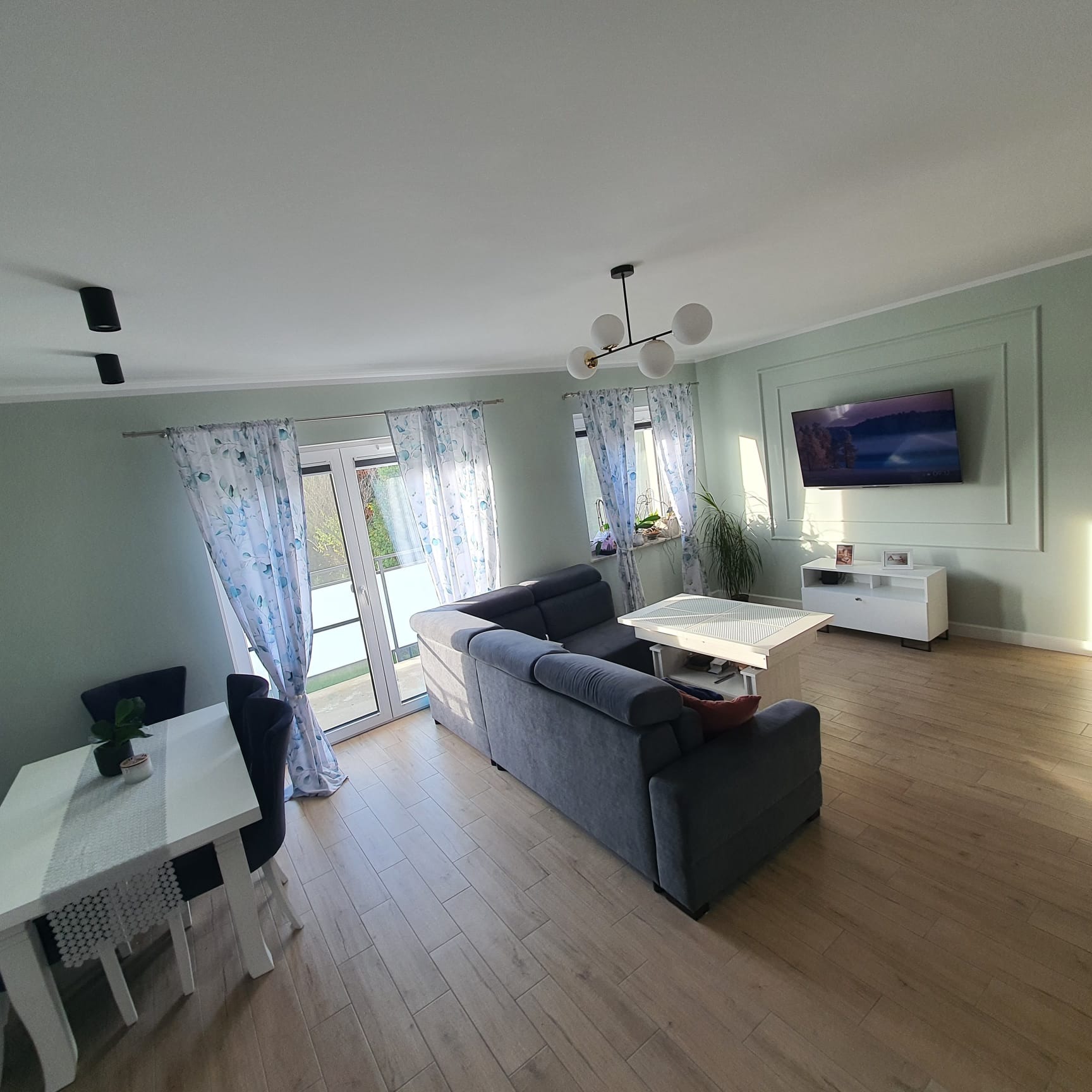 Jasne, pomalowane na zielono wnętrze salonu w Bytonii z szarym narożnikiem, białym stołem i telewizorem na ścianie z dekoracyjną ramą. Nowoczesny design.